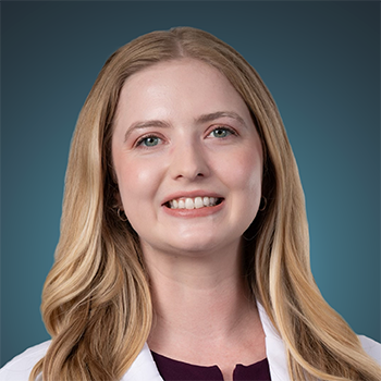 Professional woman in a white coat smiling, studio portrait with teal background, likely a healthcare provider
